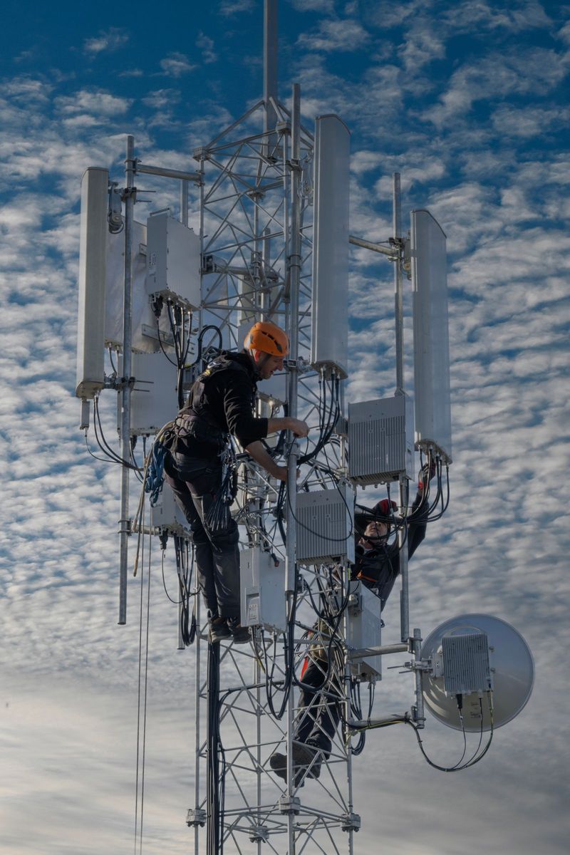 Two technicians climbing a steel telecom tower fitted with antennas and radio units